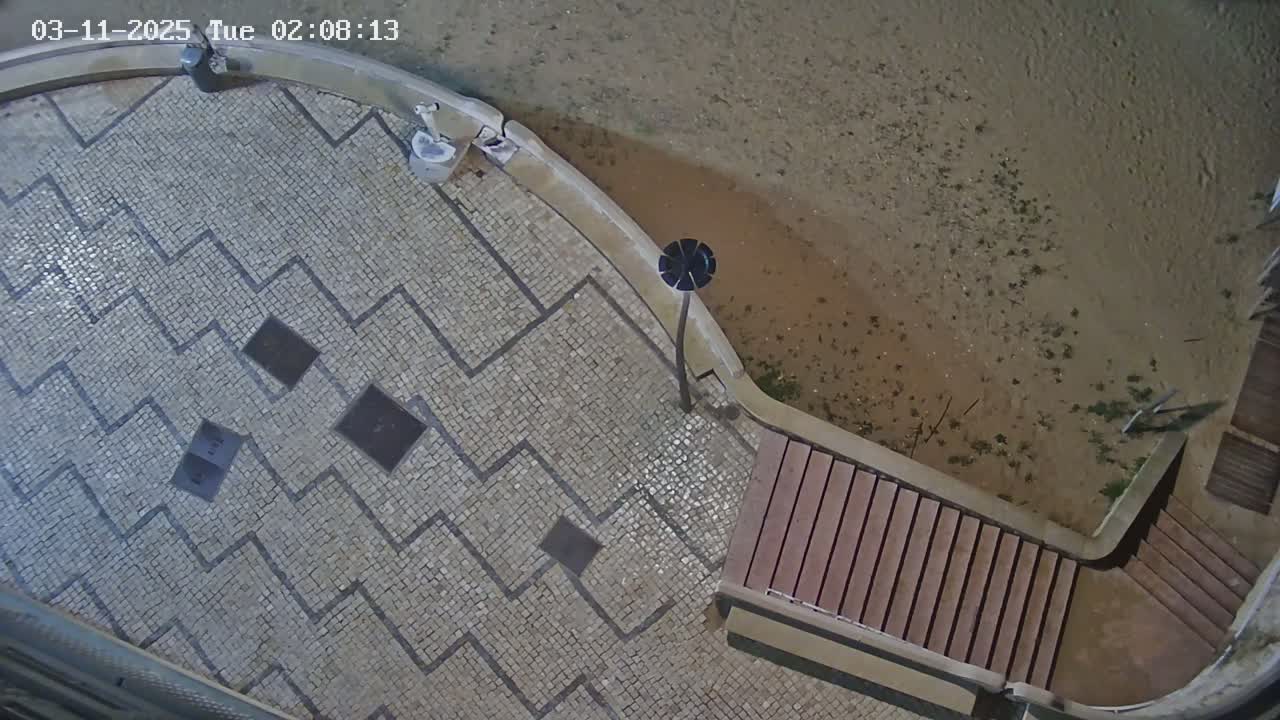 An overhead nighttime view shows a mosaic-tiled area with a zig-zag pattern, a lamppost, and wooden steps leading down to a sandy, slightly muddy area.
