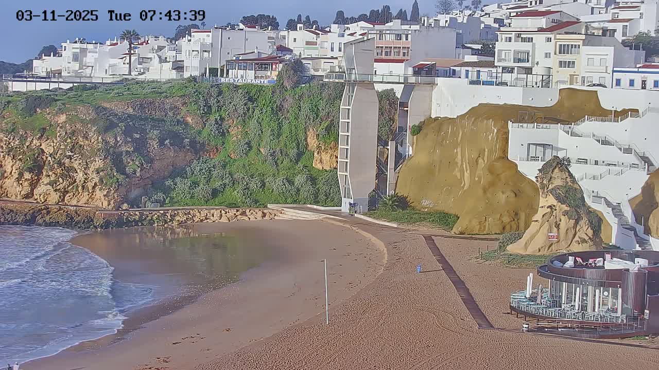A sandy beach with small waves laps against the shore below white buildings on a cliffside, under a mostly sunny sky.