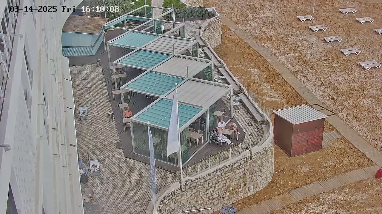 An outdoor patio area with several glass-roofed structures and seating, adjacent to a sandy beach with empty lounge chairs, and a stone wall.
