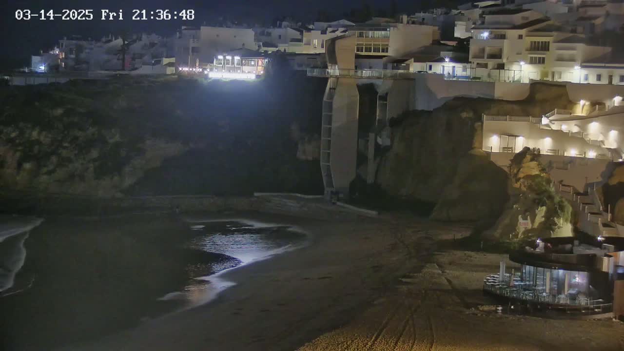 A nighttime view of a sandy beach below illuminated buildings on a cliffside.