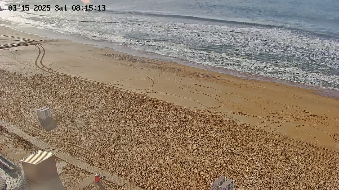 A sandy beach with tire tracks and footprints leading to a calm ocean under a mostly clear sky.