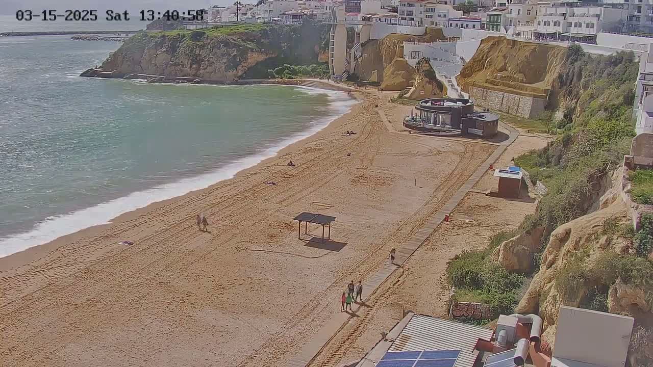A sandy beach with a few people on it, bordered by a rocky cliff and calm, light-blue ocean.