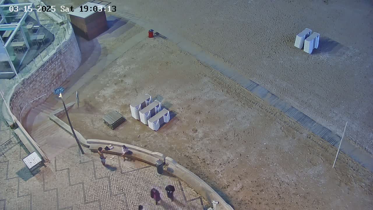 An overhead nighttime view shows a sandy beach with stacked beach chairs, a paved area with a few people, and stairs leading down to the beach.