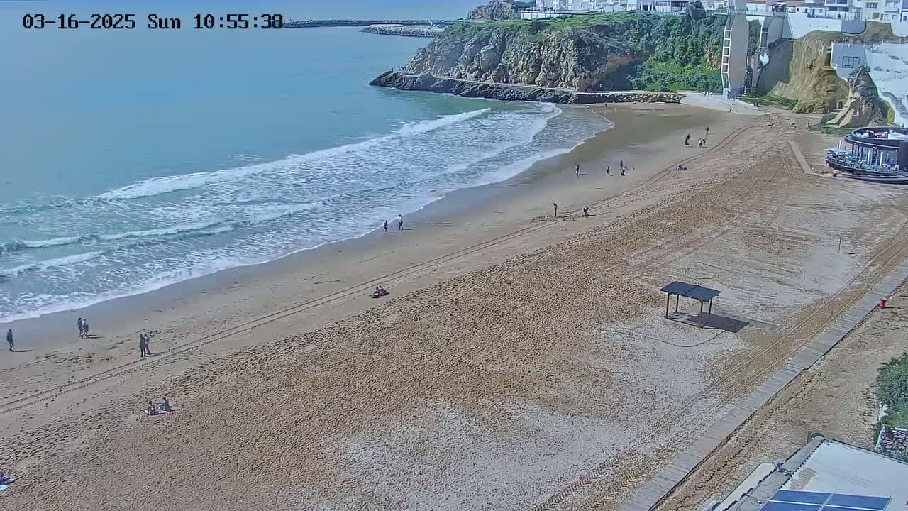 A sandy beach with a few people on it is bordered by a rocky cliff and calm ocean waves on a sunny day.