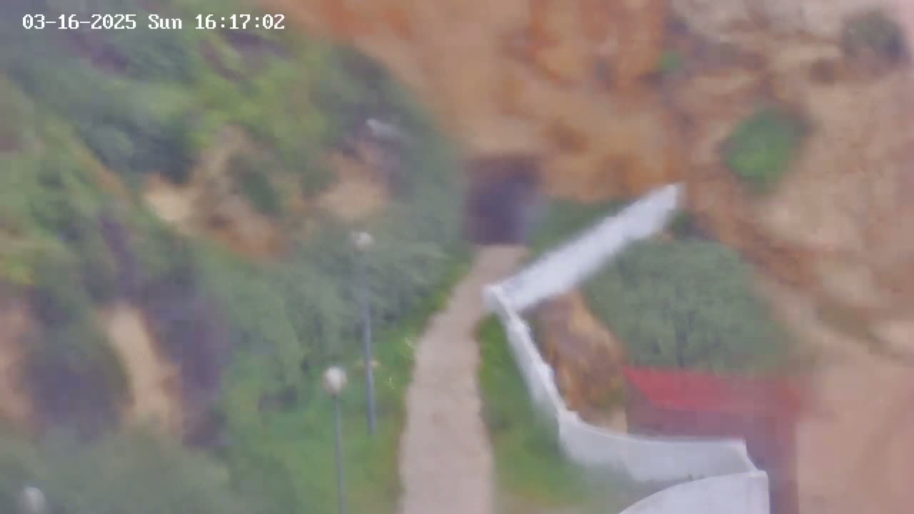 A blurry overhead view shows a pathway leading to a dark entrance, bordered by green vegetation and a white wall under an overcast sky.