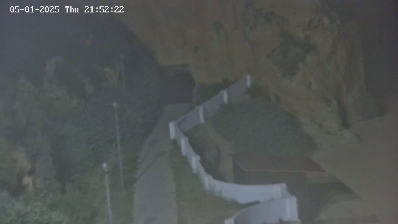 A foggy nighttime view shows a pathway alongside a white wall and rock formations, with some vegetation visible.