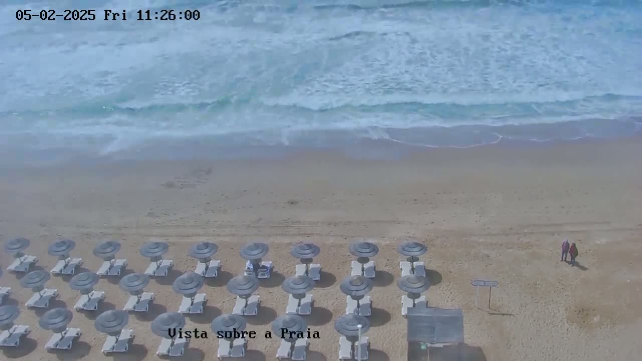 An aerial view shows a sandy beach with rows of closed umbrellas and lounge chairs, two people walking on the sand, and ocean waves rolling onto the shore.