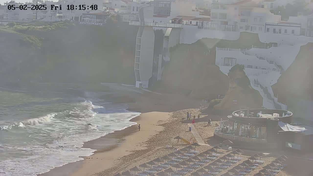 A sandy beach with gentle waves, a few people, and rows of beach umbrellas sits below white buildings on a hazy day.