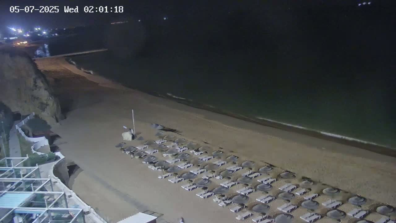 A sandy beach at night, lined with closed beach umbrellas, is gently lapped by dark water under a clear, moonless sky.