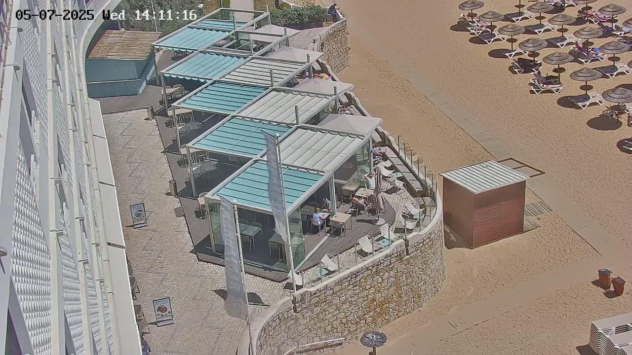 An outdoor patio restaurant with numerous covered seating areas is situated next to a sandy beach with many umbrellas and lounge chairs on a sunny day.