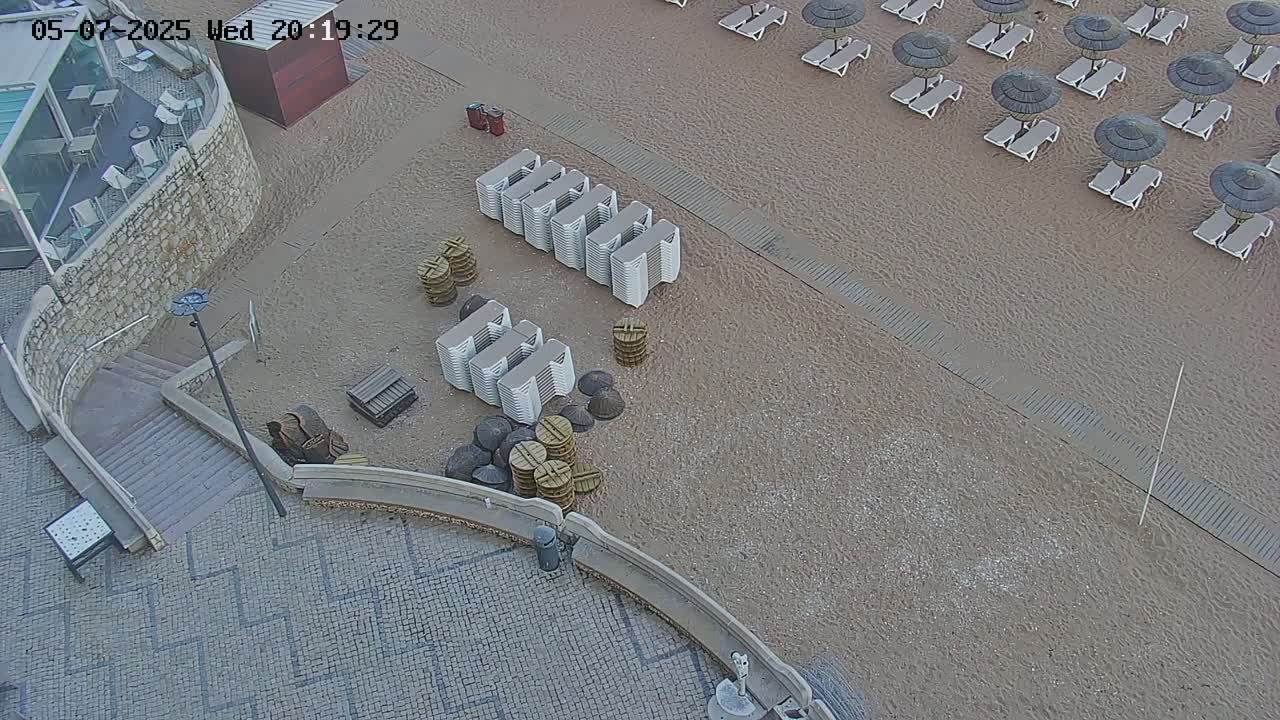 An overhead view shows a sandy beach with rows of lounge chairs and umbrellas, stacked beach chairs and other beach equipment near a stone staircase leading down to a paved area.