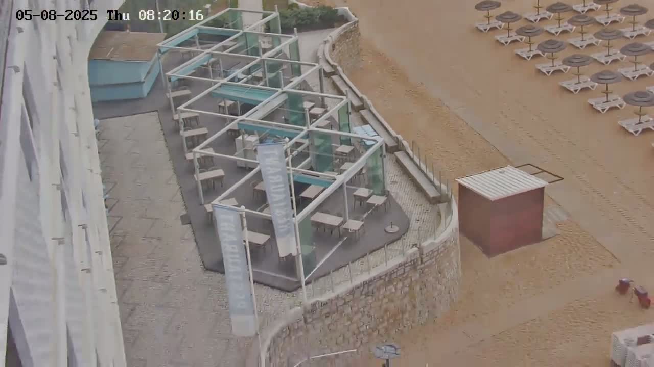 An outdoor café with glass-paneled walls and numerous tables sits adjacent to a sandy beach with rows of lounge chairs and umbrellas under an overcast sky.