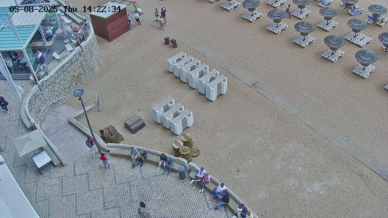 An aerial view shows a sandy beach with numerous lounge chairs and umbrellas, stacked chairs, and people sitting near a stone walkway leading to a building.