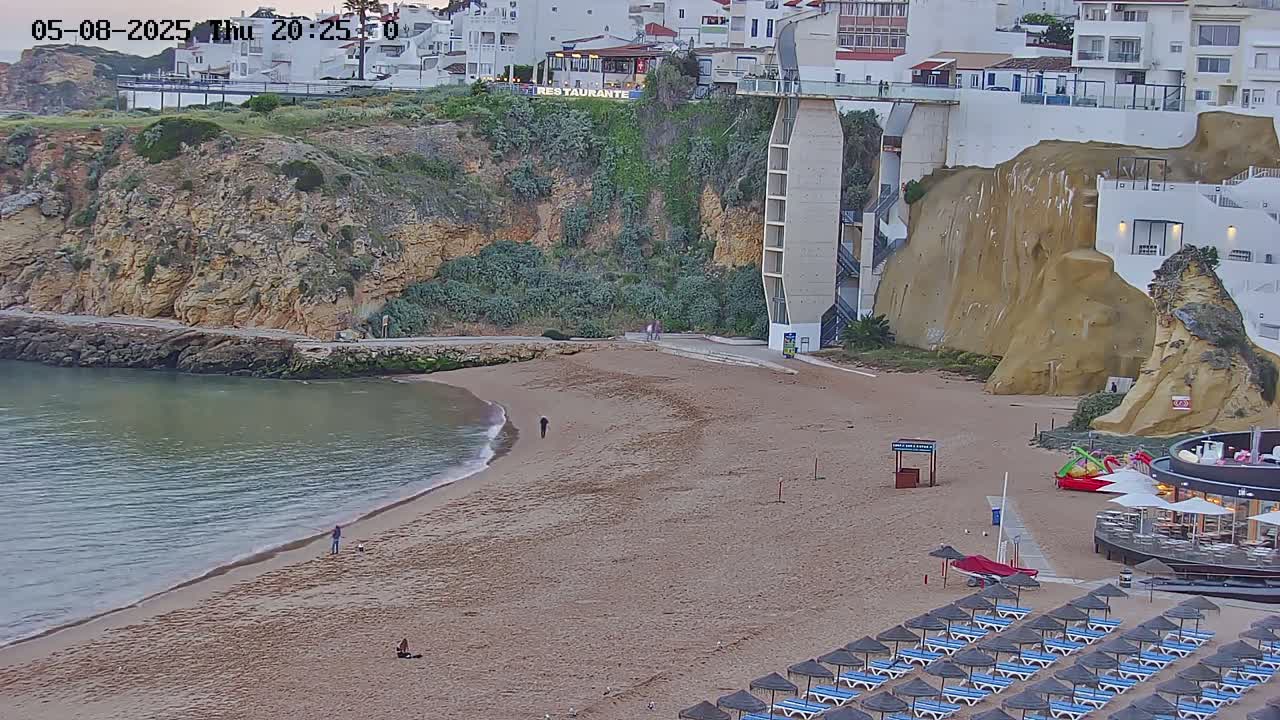 A sandy beach with calm water, a few people, and rows of closed beach umbrellas sits below a cliffside with buildings and a tall, light-colored structure.