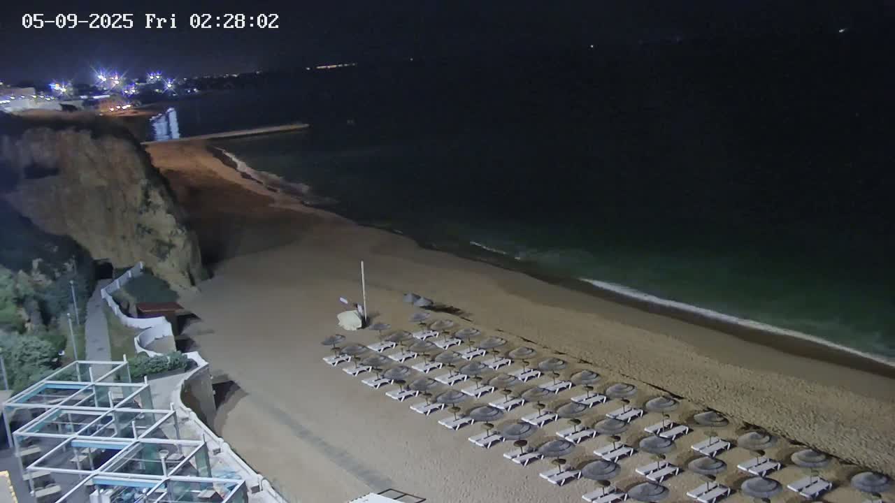 A sandy beach at night with calm waves, rows of closed umbrellas and lounge chairs, and a cliffside in the background.