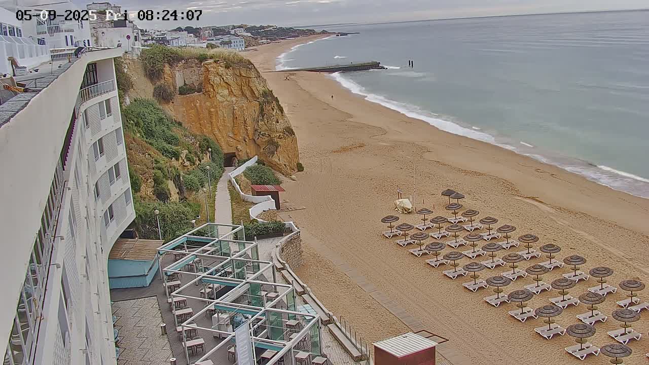 A sandy beach with rows of lounge chairs and umbrellas stretches alongside a cliff face, where a modern building is situated, all under an overcast sky.