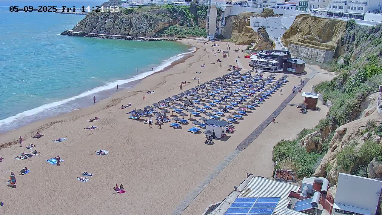 A sandy beach with numerous umbrellas and sunbathers is nestled in a cove, bordered by cliffs and calm, turquoise water under a sunny sky.