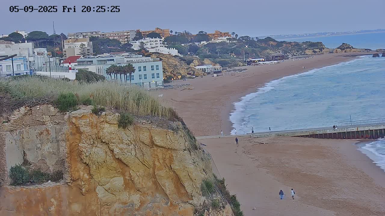 A sandy beach with gentle waves under a twilight sky is viewed from a cliff overlooking buildings and a town.