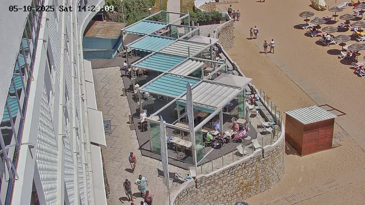 An outdoor scene on a sunny day shows a patio restaurant with multiple shaded areas and a sandy beach with people relaxing under umbrellas.