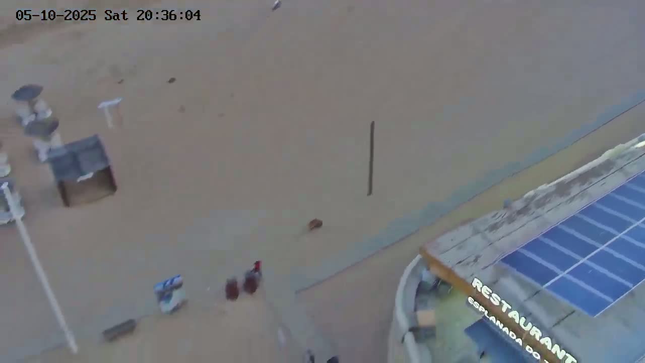 An aerial view shows a light-brown sandy area with a few small objects and a person, near a building with a solar panel on the roof.