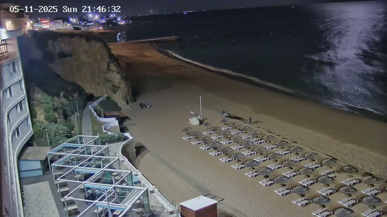 A sandy beach at night with calm ocean waves, rows of closed beach umbrellas and lounge chairs, and a cliff with a building on top.