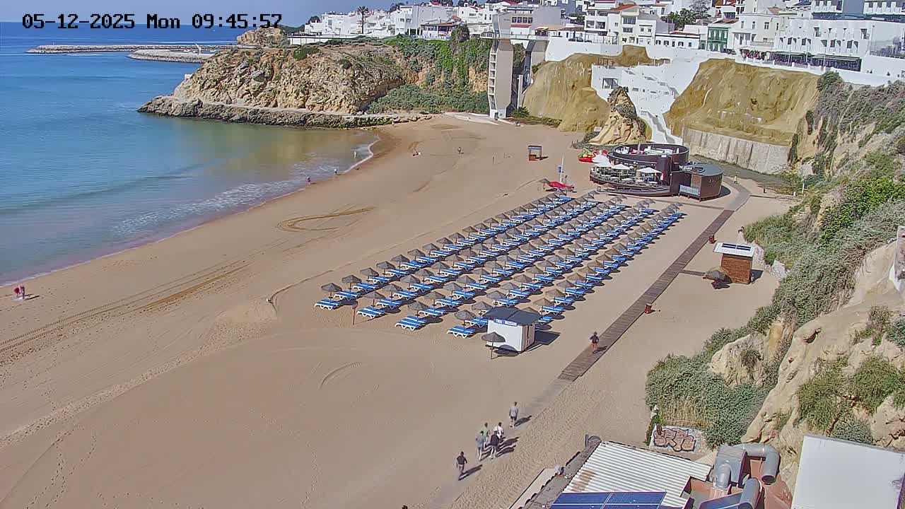 A sandy beach with rows of lounge chairs and umbrellas sits below a cliffside town on a sunny day.