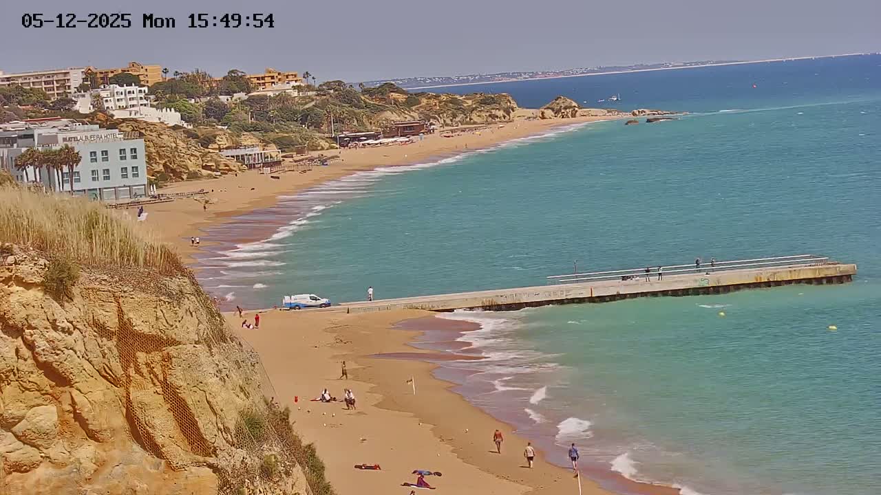 A sandy beach with turquoise water curves along a rocky cliffside under a clear sky, with a long pier extending into the sea and a few people scattered along the shore.