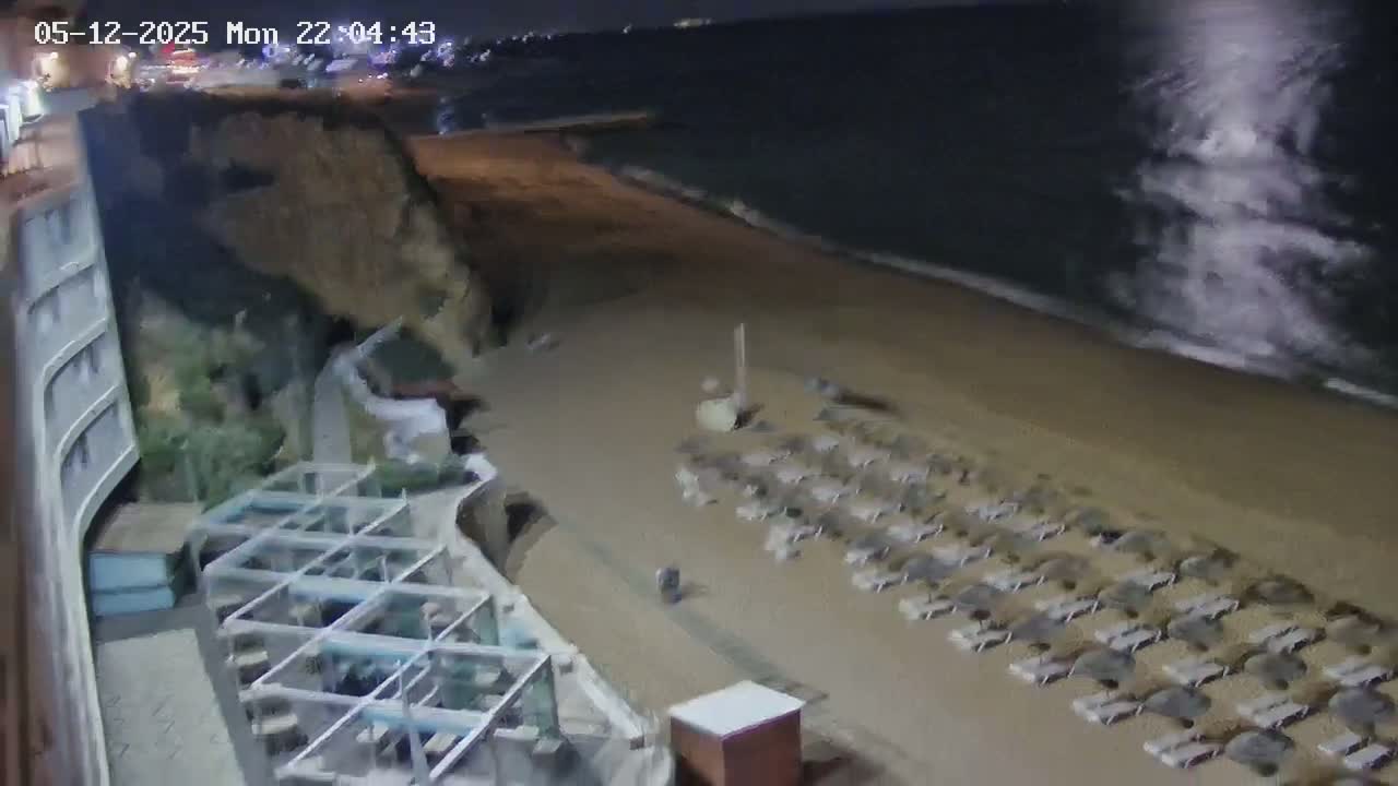 A nighttime view of a sandy beach with rows of empty lounge chairs, a building, and a dark ocean with waves gently lapping the shore.