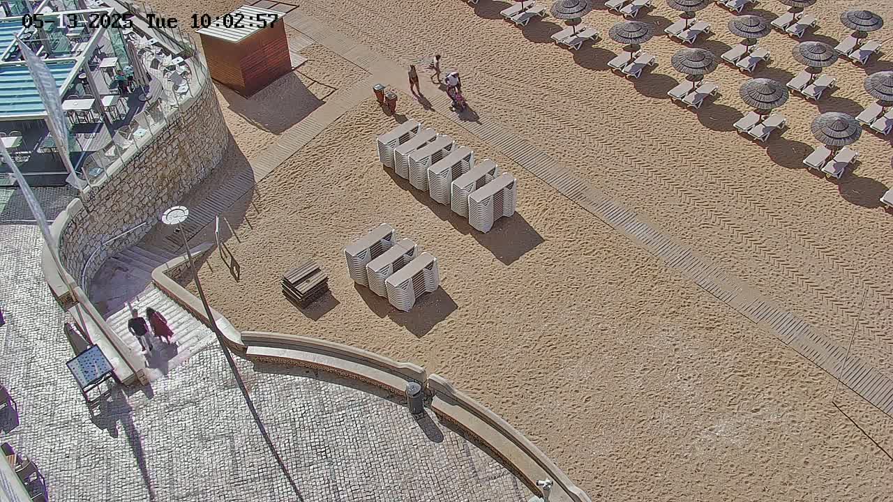 A sandy beach with numerous beach umbrellas and chairs, a stone staircase leading down from a building, and several people walking.