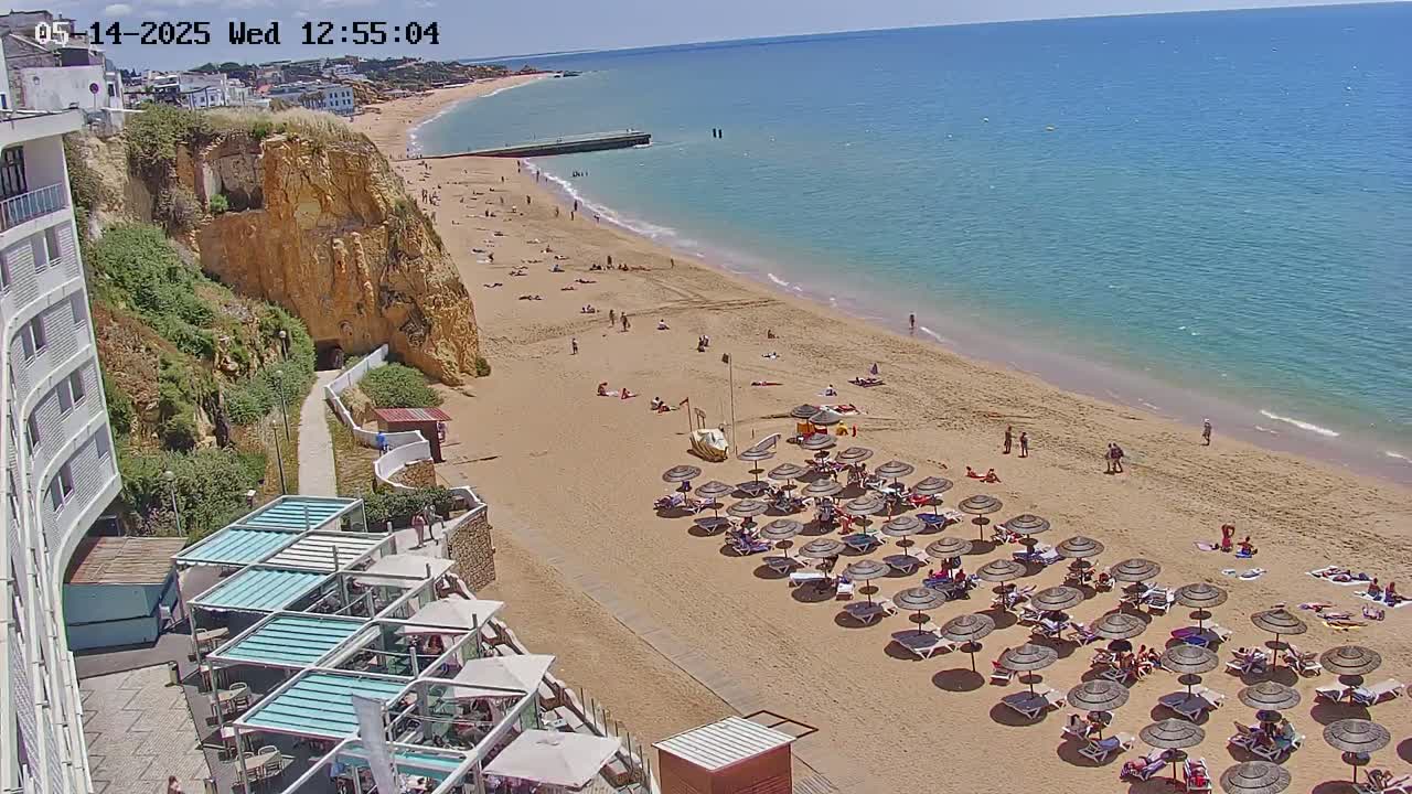 A sandy beach with numerous umbrellas and sunbathers is situated below a cliffside hotel on a sunny day, overlooking a calm, blue ocean.