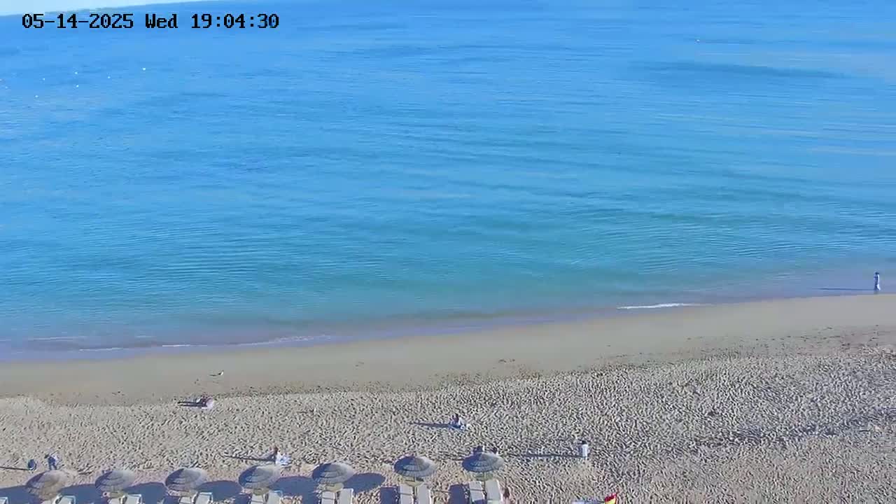 A sandy beach with calm, blue water and several closed umbrellas is visible on a sunny day.