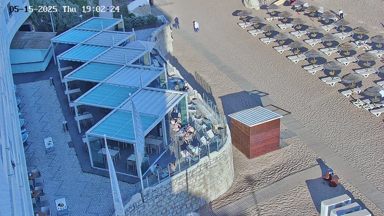 An outdoor beach scene shows a sandy beach with rows of umbrellas and lounge chairs next to a stone wall with a restaurant featuring multiple covered patio areas, all under sunny conditions.