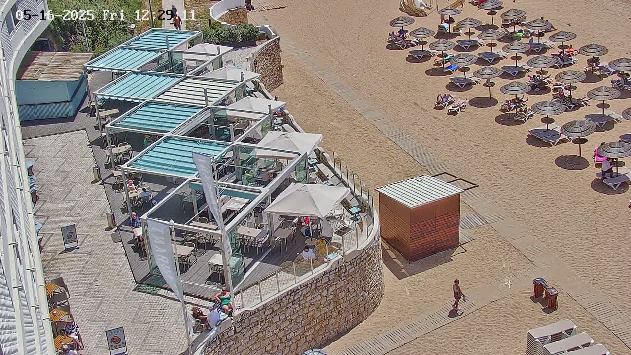 An outdoor beachside restaurant with numerous shaded tables and chairs sits adjacent to a sandy beach with many umbrellas and lounge chairs under a sunny sky.