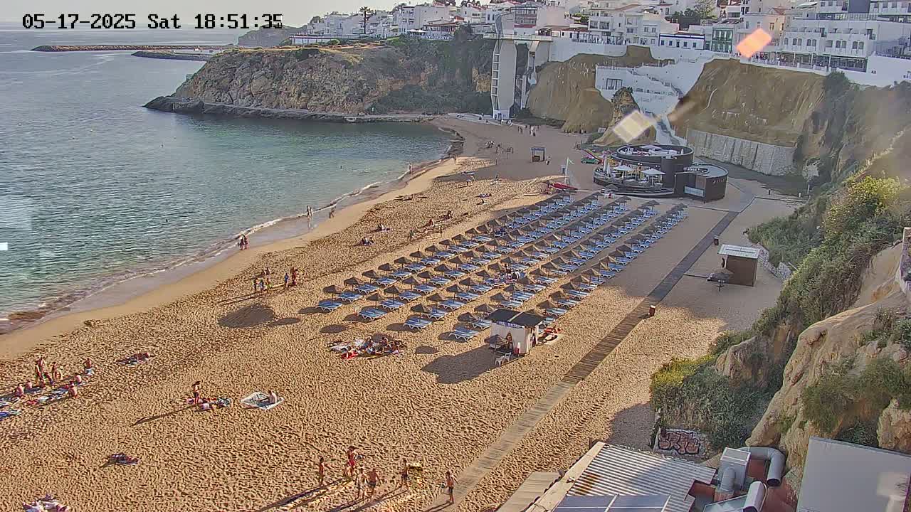 A sandy beach with numerous beach umbrellas and sunbathers is situated below a cliffside town on a sunny day.