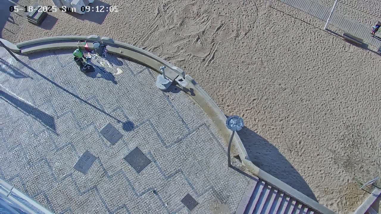An aerial view shows two people near a water fountain on a stone paved area adjacent to a sandy beach on a sunny day.