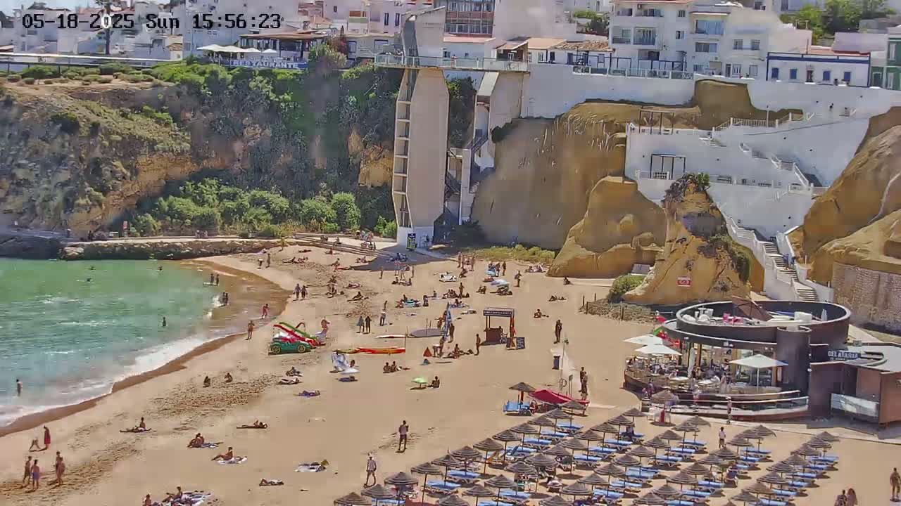A sandy beach with many people sunbathing and swimming in a clear ocean, backed by tan cliffs with white buildings and a lift, under sunny skies.