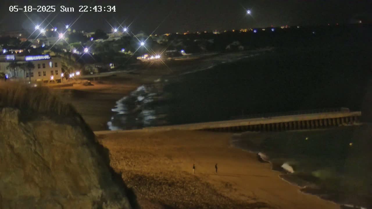 A nighttime view of a sandy beach with gentle waves, a low seawall, and a few people walking along the shore under a partly cloudy night sky.