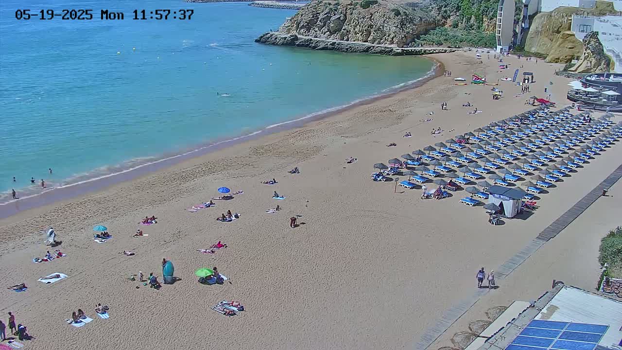 A sandy beach with turquoise water and numerous beach umbrellas and sunbathers is visible on a sunny day.
