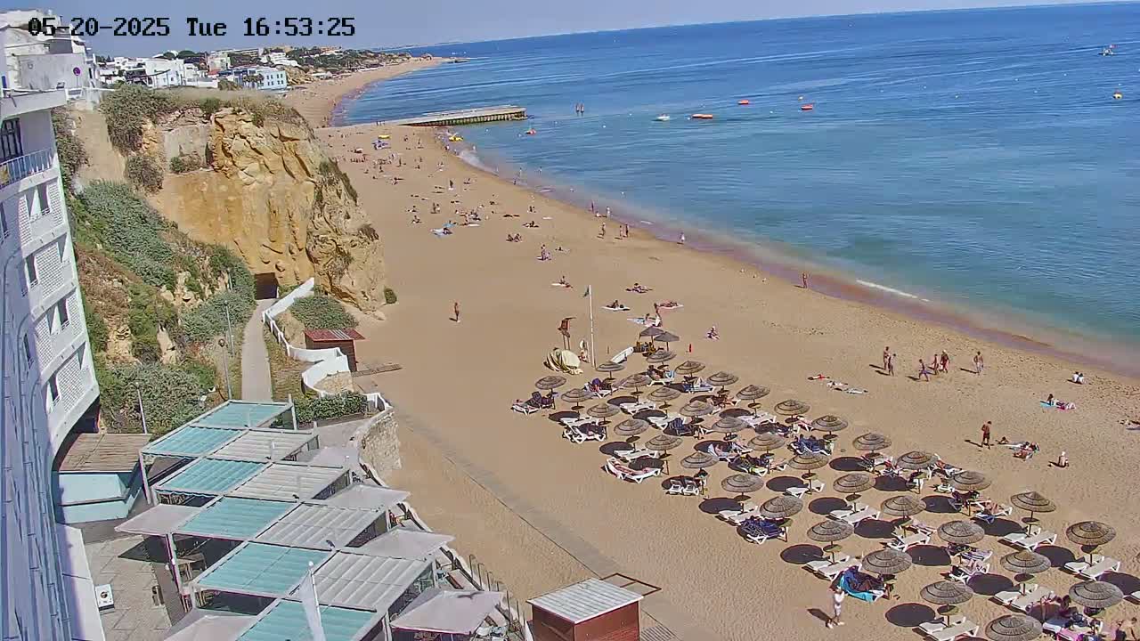 A sandy beach with numerous beach umbrellas and sunbathers is situated below a cliff next to a calm blue ocean on a sunny day.