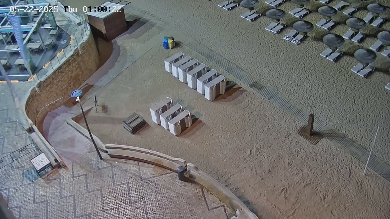 At night, a sandy beach area with folded beach chairs and umbrellas is seen from above, next to a paved walkway and a building with outdoor seating.