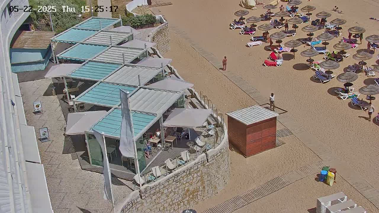 An outdoor beach scene on a sunny day shows a restaurant with multiple covered patio areas next to a sandy beach with numerous umbrellas and beachgoers.