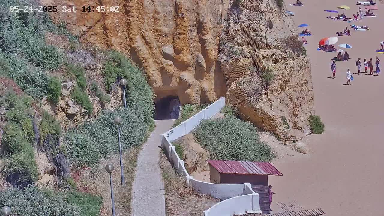 A paved walkway leads to a tunnel carved into a sandy cliff overlooking a sunny beach with people sunbathing and swimming.