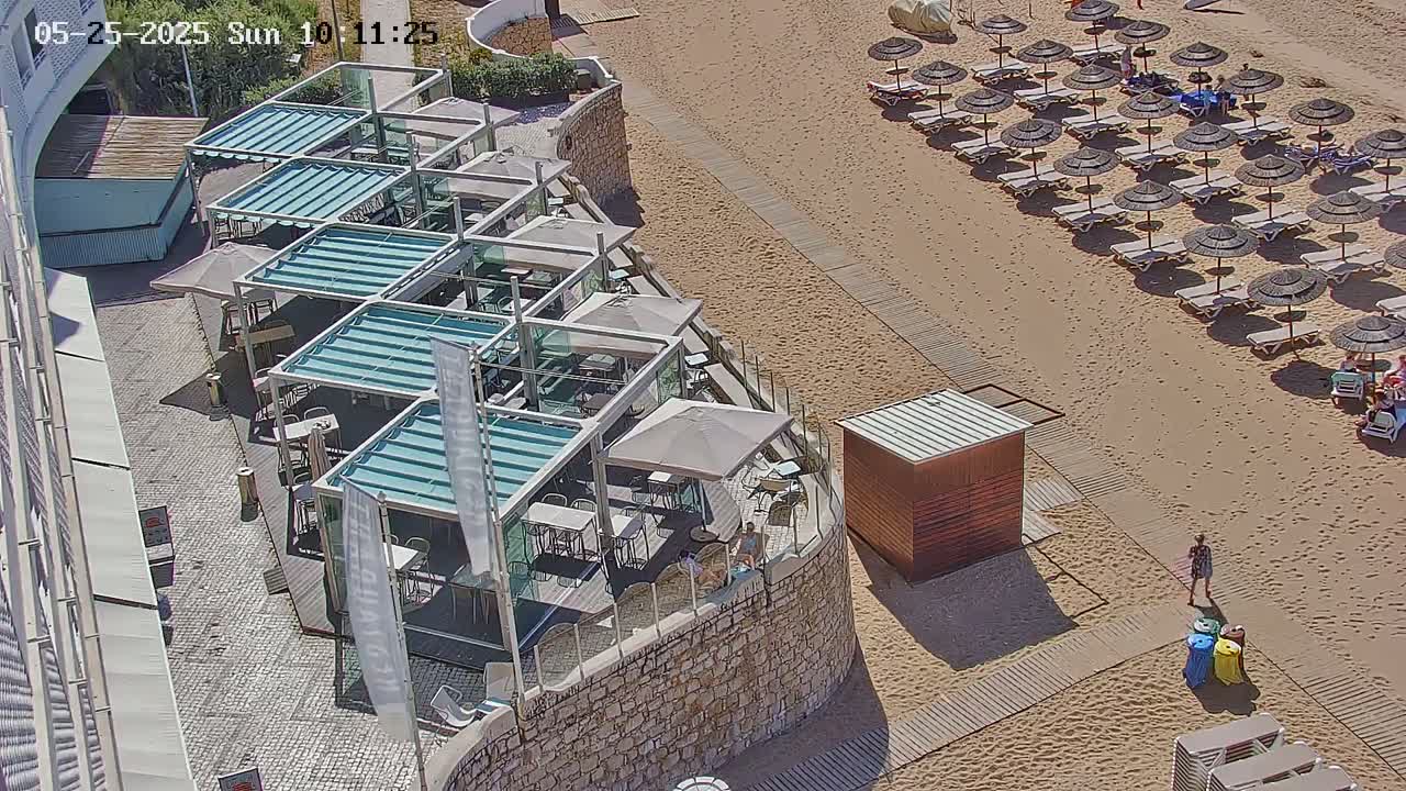An outdoor cafe with numerous shaded tables and chairs is situated next to a sandy beach with rows of umbrellas and lounge chairs under a sunny sky.