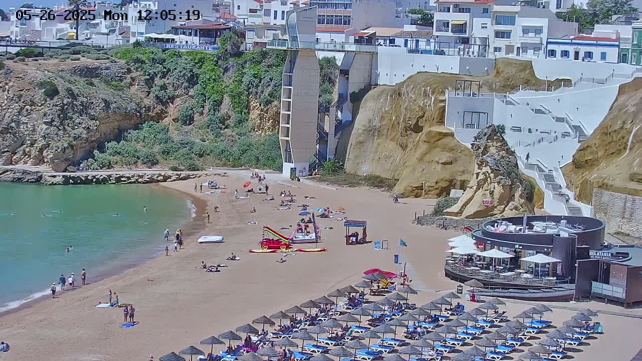 A sandy beach with people swimming and sunbathing, lined with lounge chairs and umbrellas, sits below a cliffside with buildings and a stairway on a sunny day.