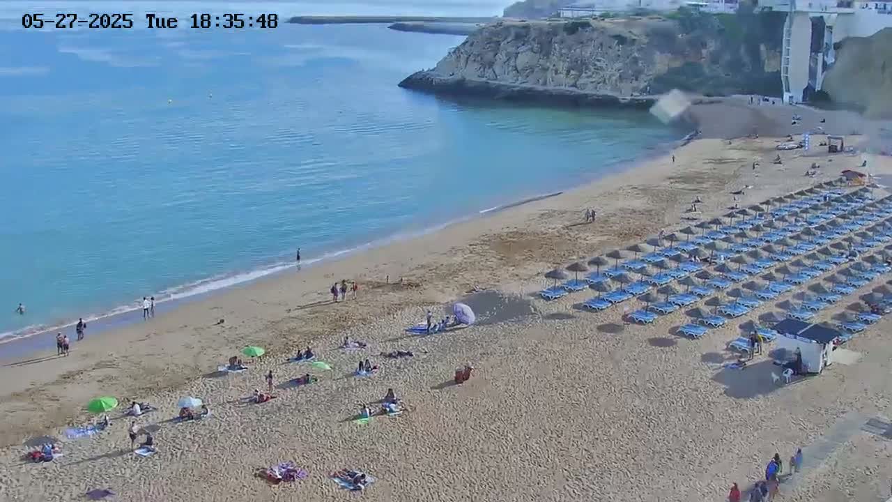 A sandy beach with numerous blue umbrellas and lounge chairs is partially populated with people on a sunny day, next to a calm blue ocean with a rocky cliff in the background.