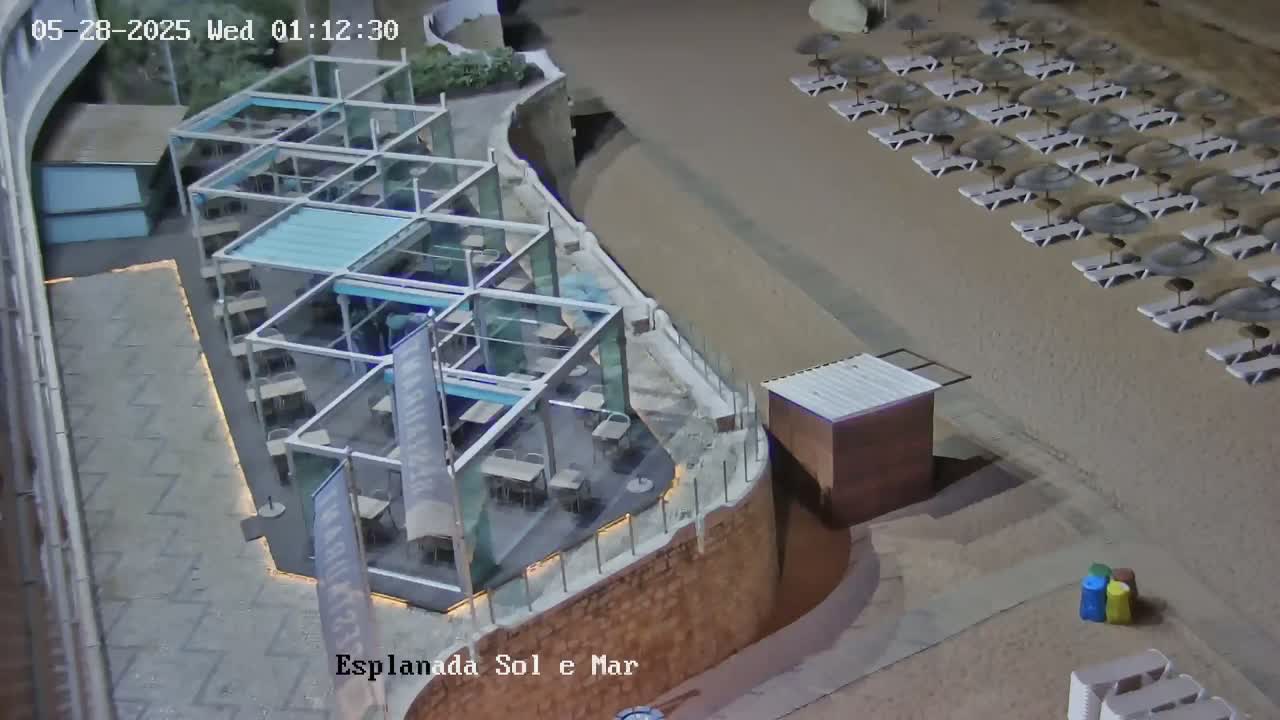 A nighttime aerial view shows a beach with rows of empty lounge chairs and umbrellas next to an outdoor restaurant with a glass-roofed structure.