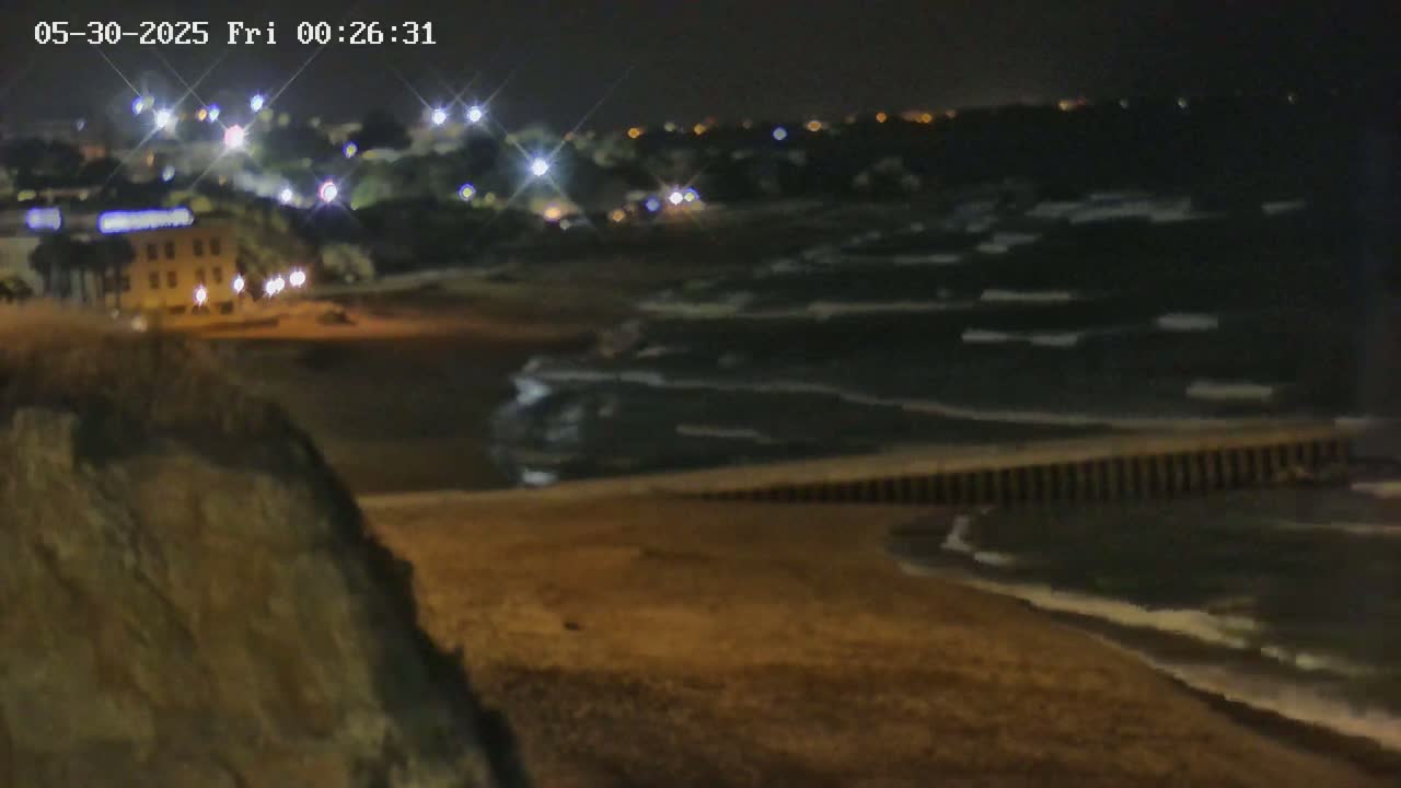 A nighttime view of a dark, wavy ocean meeting a sandy beach, with a town's lights visible in the distance.