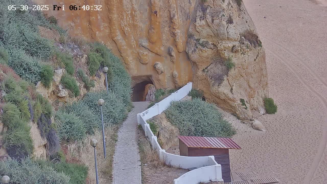 A paved pathway leads to a tunnel carved into a tan cliff overlooking a sandy beach.