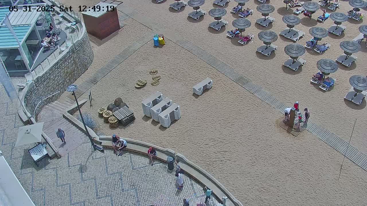An aerial view shows a sandy beach with numerous lounge chairs and umbrellas under a sunny sky, adjacent to a paved area with people and some buildings.