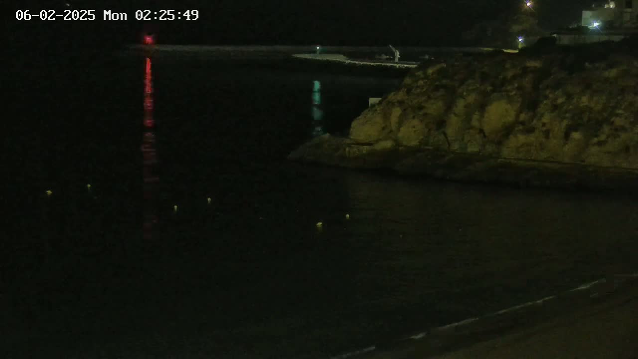 A nighttime view of dark water reflecting red and green lights, with a rocky shoreline visible under a clear, dark sky.